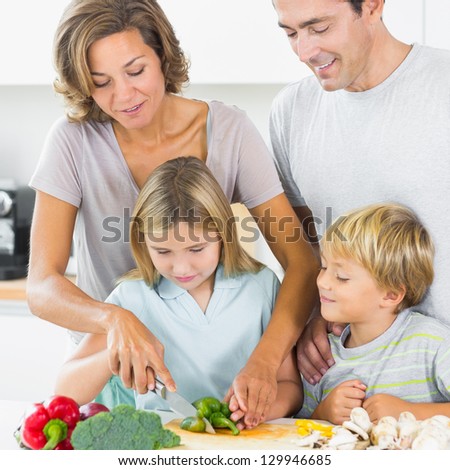 Mother teaching daughter to slice vegetables as father and son are watching in the kitchen