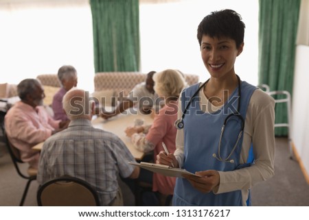 Front view of female doctor holding clipboard and pen with group of senior people playing card behind in living room at nursing home