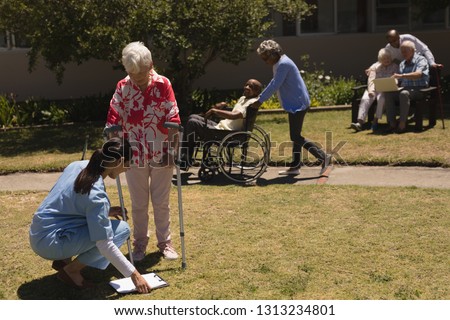Front view of young female doctor kneeling and examining in front of disabled senior woman in garden on a sunny day