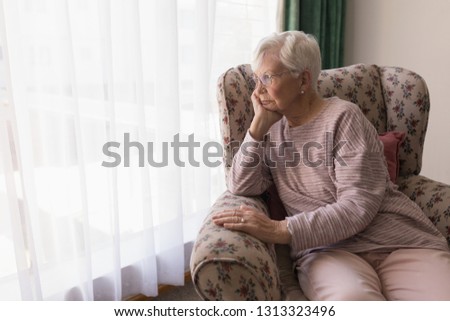 Front view of thoughtful senior woman sitting on the couch and looking outside through window at nursing home
