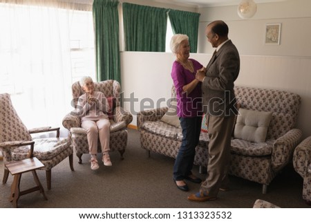 Side view of happy senior friends dancing together with their friend sitting on the sofa at nursing home