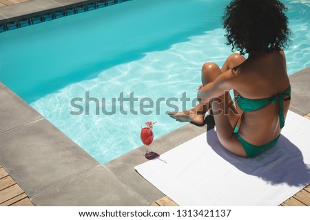 Rear view of beautiful young African American woman sitting at poolside in her backyard on a sunny day