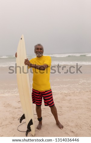 Front view of happy senior male surfer standing with surfboard and looking at camera on the beach