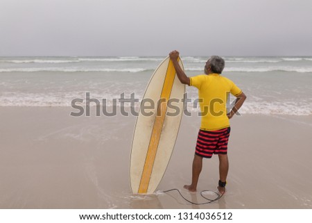 Rear view of active senior male surfer standing with surfboard on the beach