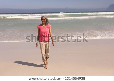 Front view of happy senior woman looking at camera while walking on beach in the sunshine
