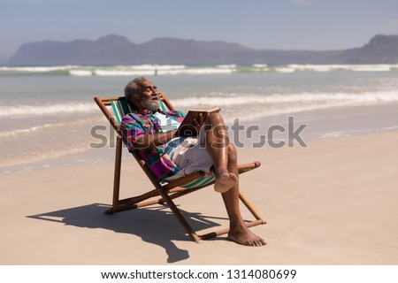 Side view of senior man relaxing on sun lounger and reading a book on beach in the sunshine