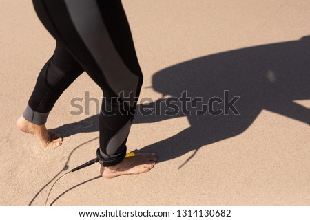 Low section of senior woman walking with surfboard leash on her leg on the sand at beach