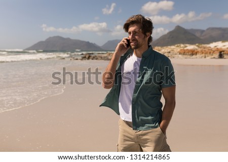 Front view of young man with hand in pocket talking on mobile phone on beach in the sunshine