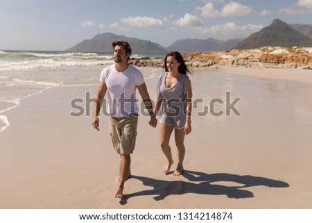 Front view of happy young couple holding hands and walking on beach in the sunshine