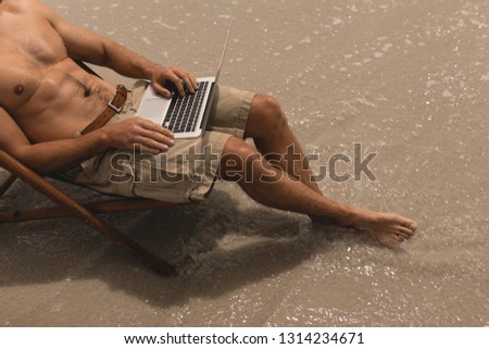 Low section of shirtless young man relaxing on sun lounger and using laptop on beach in the sunshine