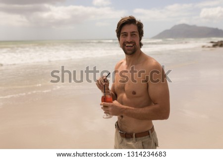 Front view of shirtless happy young man with cocktail looking at camera on beach in the sunshine