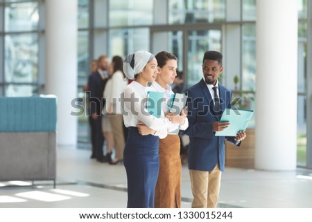 Side view of young business executives standing with files in lobby office against diverse business people interacting in background