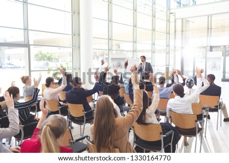 Rear view of interactive diverse business people listening to Caucasian businessman and raising hands to ask him questions in conference