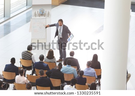 High angle view of well dressed Caucasian businessman speaking and holding digital tablet and microphone while pointing out something on flip chart to group of diverse business people at conference