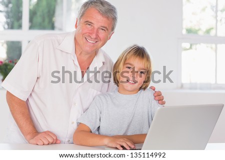 Grandfather and child looking at the camera with smile in front of laptop in kitchen