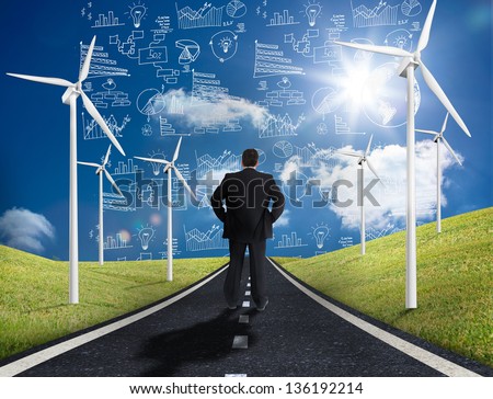 Businessman standing in middle of road with wind turbines either side looking at graphs on the horizon