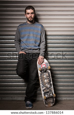 Attractive skater leaning against metal shutters and holding his board in the skate park