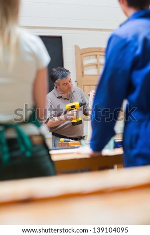 Teacher in a woodwork class holding a wooden board while explaining and drilling