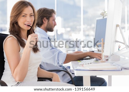 Woman biting her glasses with colleague working behind in creative office