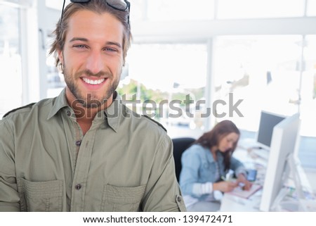 Cheerful man in creative office with arms folded and partner working behind
