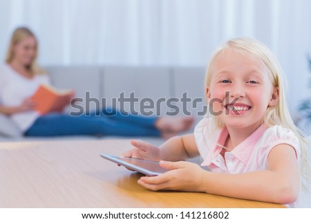 Little girl using tablet pc in the living room while her mother is reading a book