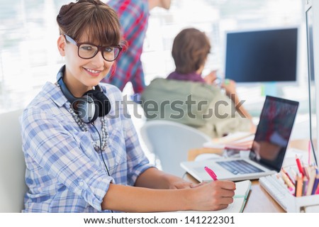 Portrait of an attractive designer writing on a notepad with colleagues discussing behind