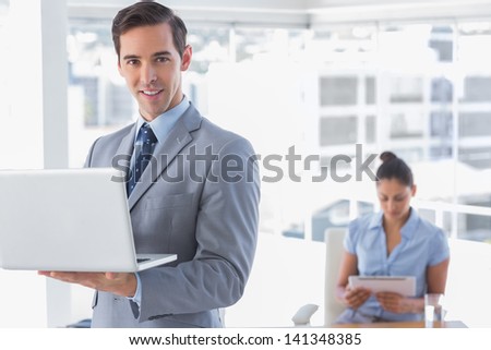Businessman standing with laptop and smiling at camera with woman working behind him