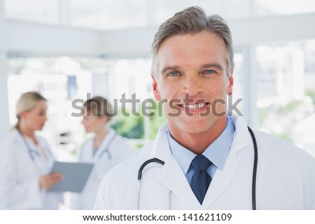 Smiling grey haired doctor standing in his office with colleagues working behind