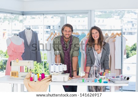 Smiling fashion designers leaning on desk with spools of thread and sewing machine