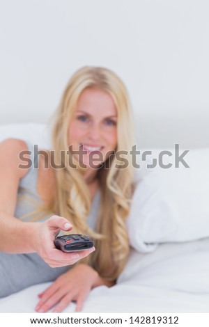 Blonde woman pointing the remote control at the camera in her bed
