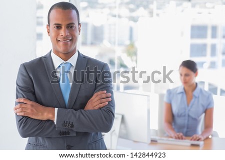 Businessman standing with arms crossed with woman working behind