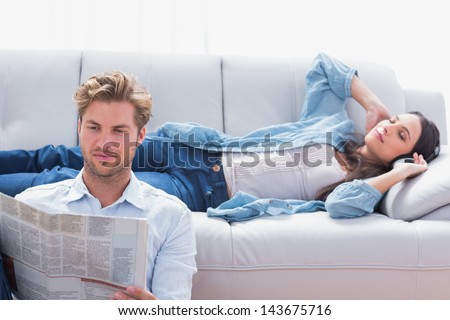 Woman laid on a couch listening to music next to her boyfriend reading a newspaper