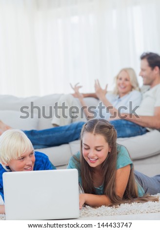 Siblings using a laptop lying on a carpet with parents discussing behind