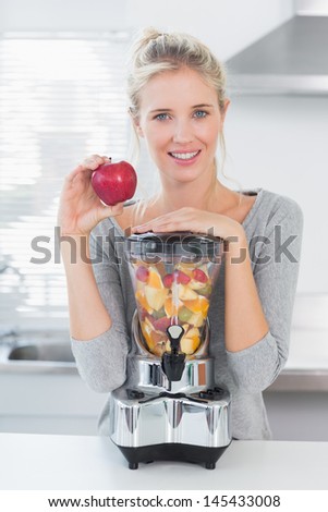 Pretty woman leaning on her juicer full of fruit and holding red apple at home in kitchen