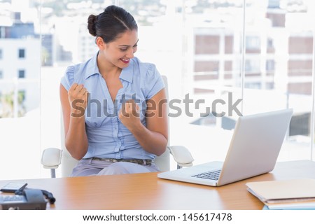 Brunette businesswoman cheering in front of her laptop at her desk in the office