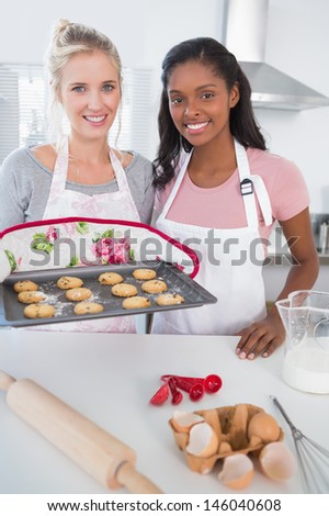 Smiling woman showing freshly baked cookies with friend at home looking at camera in kitchen