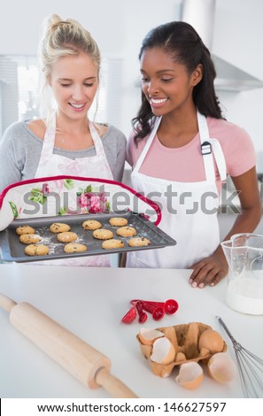 Smiling woman showing freshly baked cookies to friend at home in kitchen