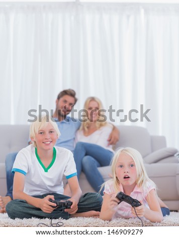 Children on the carpet playing video games in the living room while their parents are watching them