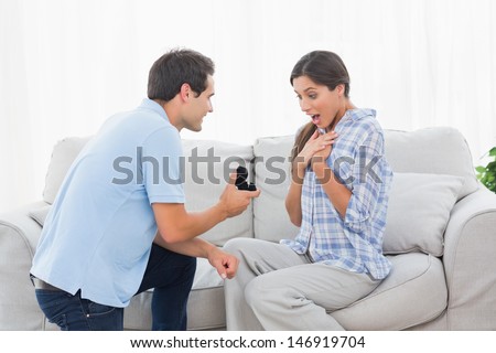 Man on bended knee offering an engagement ring to his partner on the couch