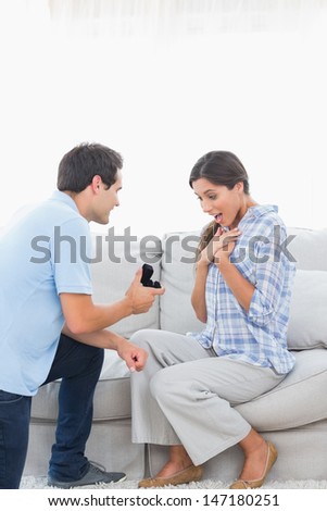 Man on bended knee offering an engagement ring to his girlfriend on the couch
