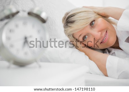 Blonde woman covering her ears from alarm clock noise at home in bed
