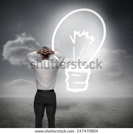 Businessman looking at light bulb with hands on head in dark grey landscape