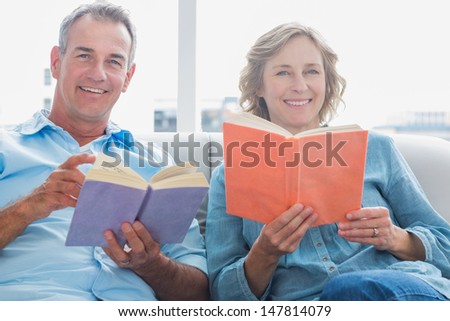 Relaxed couple reading books on the couch smiling at camera at home in the living room