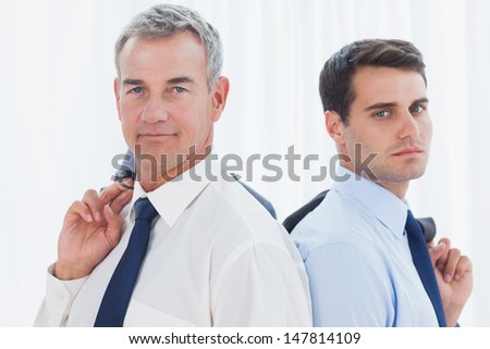 Serious businessmen in bright office posing back to back together while holding their jacket