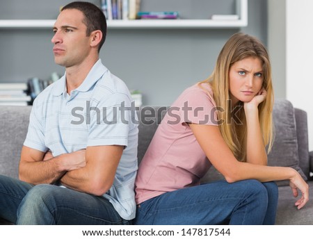 Couple sitting back to back after a fight on the couch with woman looking at camera in sitting room at home