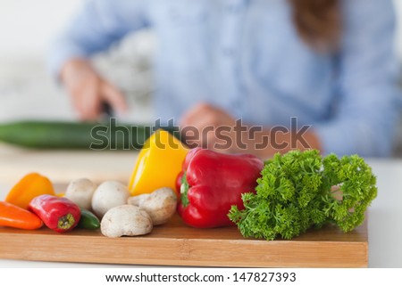 Wooden board with vegetables on a table with a woman cutting a courgette on the background