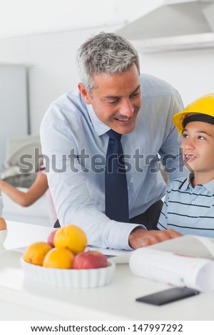 Father showing son his blueprints as he is wearing yellow helmet in the kitchen before work