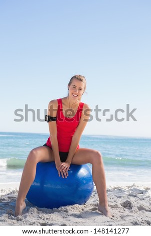 Fit blonde sitting on exercise ball smiling at camera on the beach