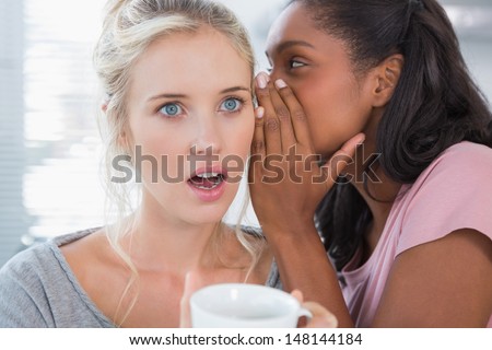 Young woman whispering secret to her shocked friend at home