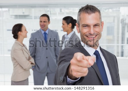Cheerful businessman in bright office pointing at camera with colleagues on background